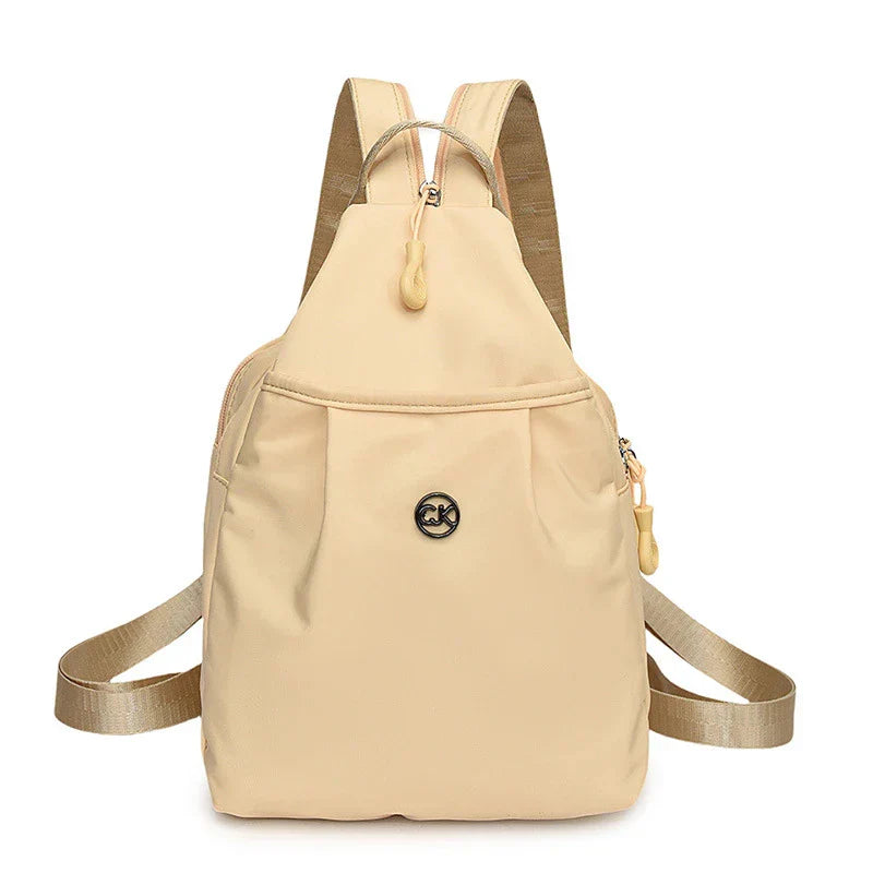 Bolso convertible de hombro: vista frontal de una mochila pequeña color beige claro con correas para los hombros, asa superior y insignia central con logotipo