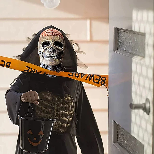 halloween kostume - Person i kraniemaske og sort kappe, der holder en sort jack-o-lantern-spand, stående ved en dør bag omvendt orange advarselstape.