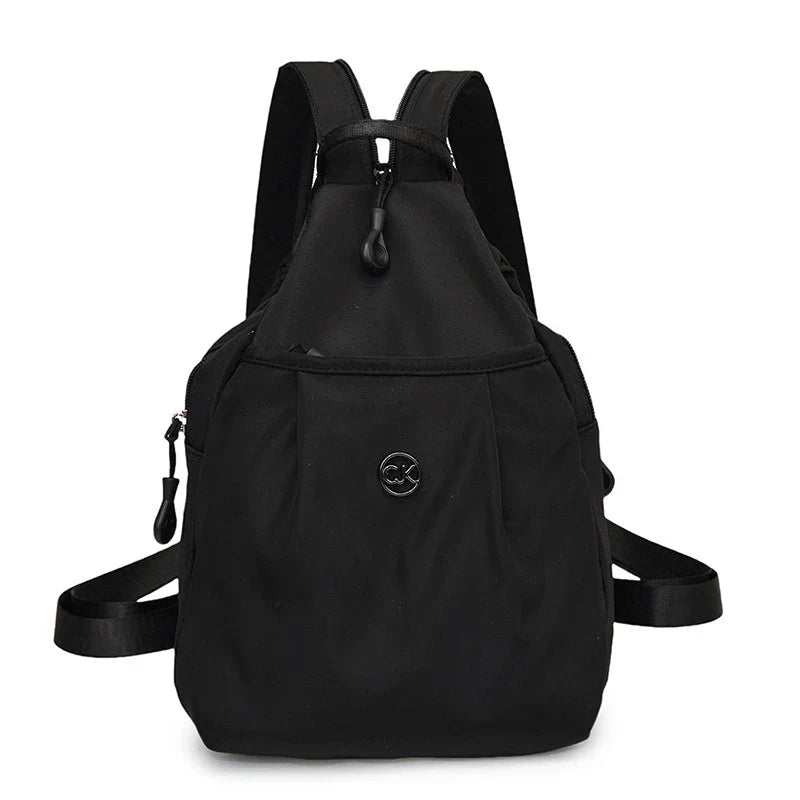 Bolso convertible de hombro: mochila compacta negra sólida con correas dobles para los hombros, tiradores de cremallera y un pequeño emblema circular frontal, vista desde el frente sobre un fondo blanco
