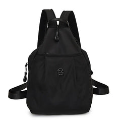 Bolso convertible de hombro: mochila compacta negra sólida con correas dobles para los hombros, tiradores de cremallera y un pequeño emblema circular frontal, vista desde el frente sobre un fondo blanco