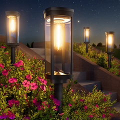 Solar Pathway Lights - Black cylindrical stake light with warm filament-style glow installed among pink flowering shrubs along a sloped stone staircase at night
