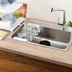 Expandable rolling utility cart placed across a kitchen sink holding inverted glasses and a cup, with a dish rack nearby on the countertop