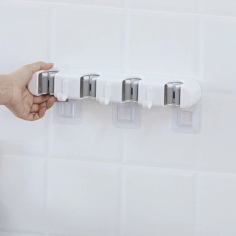 soporte de pared para trapeador y escoba - Mano sosteniendo un organizador de pared blanco con 4 ranuras junto a tres almohadillas adhesivas transparentes en una pared de azulejos blancos