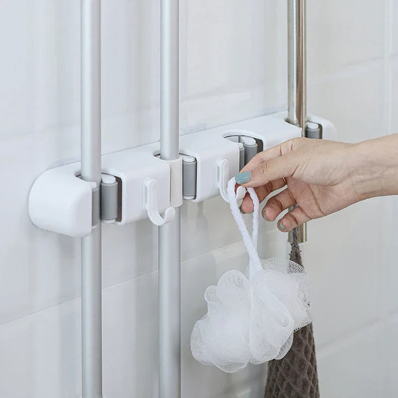soporte de pared para trapeador y escoba - Mano colgando una esponja de baño blanca en un gancho de un soporte de pared para herramientas que sostiene tres herramientas con mango plateado