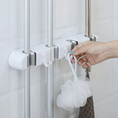 soporte de pared para trapeador y escoba - Mano colgando una esponja de baño blanca en un gancho de un soporte de pared para herramientas que sostiene tres herramientas con mango plateado