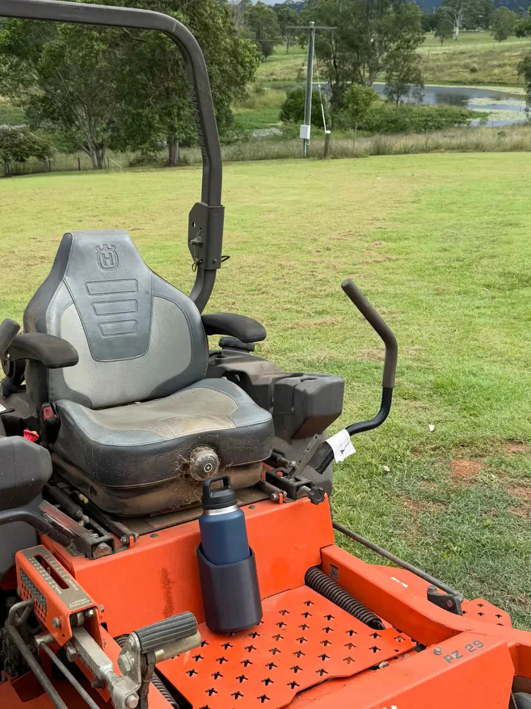 Water Bottle Holder - Dark blue cup holder holding a blue water bottle, mounted on an orange zero-turn mower footrest on grass