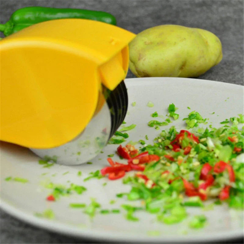 Pizza Dough Cutter Slicer - Yellow handheld slicer resting on a white plate with chopped green herbs and red chili pieces, with a potato and pepper in the background.