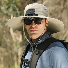 sombrero de ala ancha - Persona usando un sombrero de ala ancha beige claro con un ventilador solar en el ala frontal, gafas de sol y mochila visibles