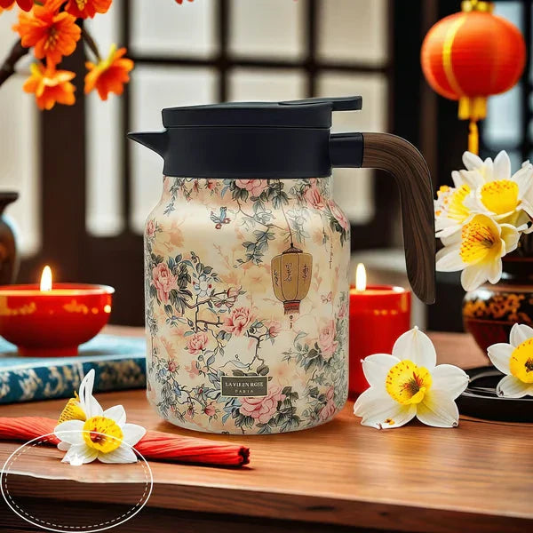 Vintage Floral Tea Thermos - Insulated pitcher with light beige traditional floral and lantern print, black lid, wood-grain handle, on a wooden table with candles and daffodils
