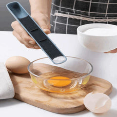 Hand holds an adjustable measuring spoon pouring white powder into a glass bowl with a raw egg, on a wood cutting board