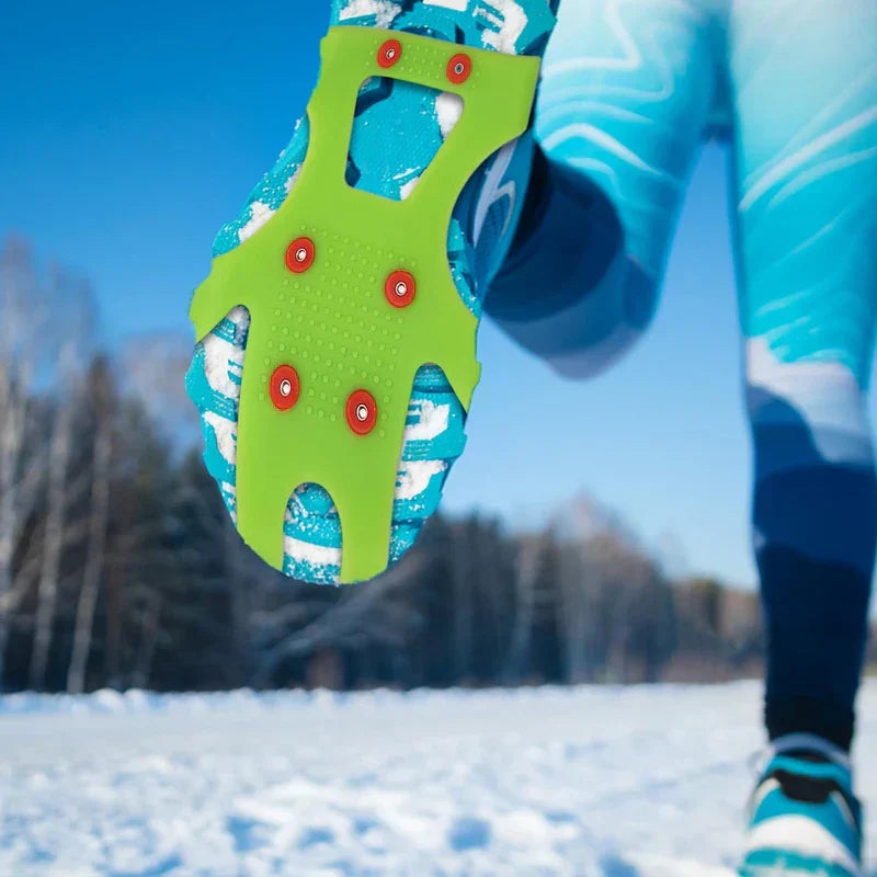 Close-up of green ice cleats crampons fitted to a blue running shoe sole, worn by a person on a snow-covered outdoor path.