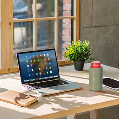 portavasos aislante para latas con base antideslizante - portavasos verde oliva sosteniendo una lata de refresco roja, sobre un escritorio iluminado por el sol junto a un portátil, cuaderno, planta en maceta y tableta junto a una ventana