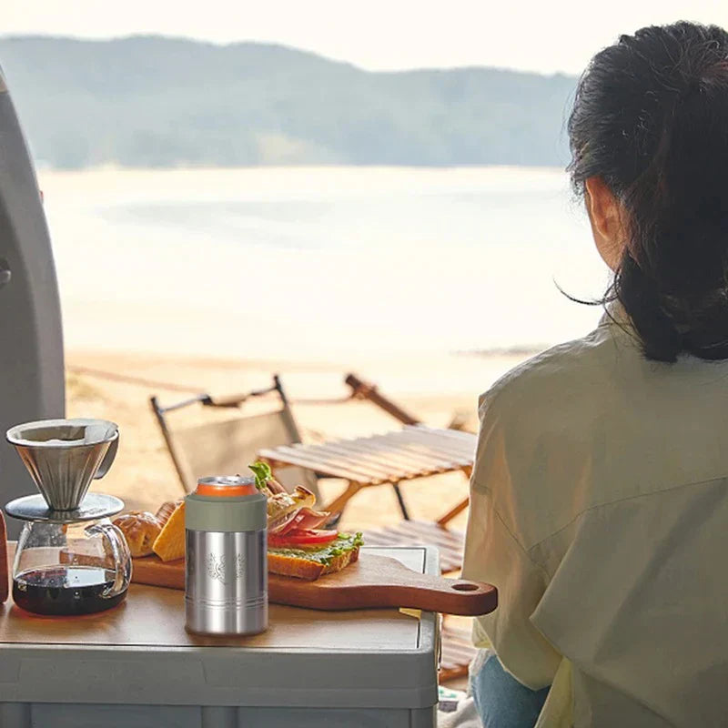 portavasos aislante para latas con base antideslizante - portavasos plateado con parte superior verde oliva sosteniendo una lata naranja, sobre una mesa de campamento con equipo de café y comida, con vista a una playa junto al lago