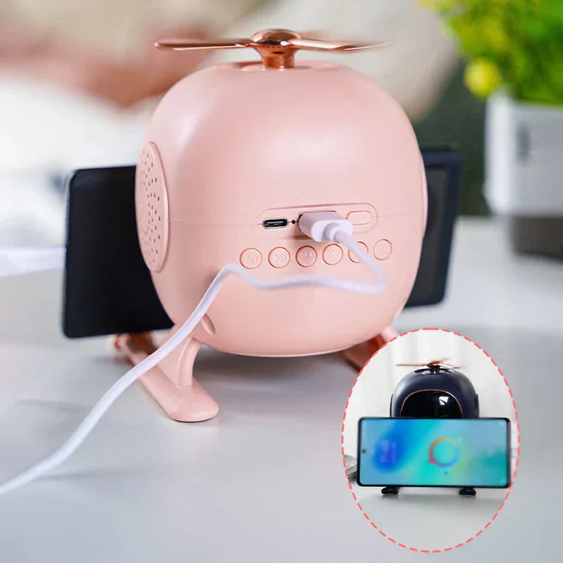 Pink helicopter-shaped bluetooth speaker with charging cable plugged in, shown with phone stand function