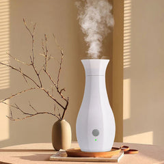 Máquina de perfume - Humidificador blanco con forma de jarrón emitiendo niebla, sobre una bandeja encima de una revista, junto a un pequeño florero con ramas secas, sobre una mesa clara con sombras de pared rayadas