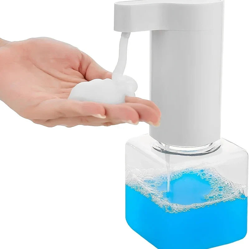 A hand under a white automatic soap dispenser receiving foamy soap from a clear bottle with blue liquid.