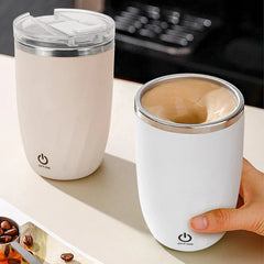 Automatic Mixing Mug Cup - Two self-stirring cups on a desk: a standing light pink lidded cup, and a held white cup with swirling brown liquid inside