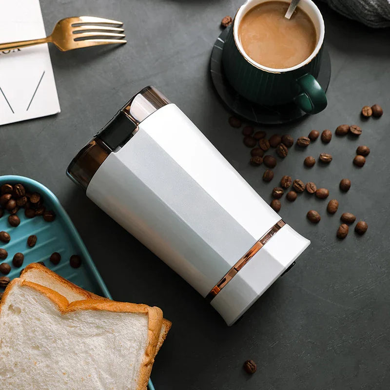 Coffee Grinding Machine - White and rose gold electric grinder lies on a dark table, surrounded by coffee beans, sliced bread, a green coffee cup, and a golden fork