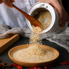Coffee Grinding Machine - A person holds a tilted white grinder, using a wooden spoon to scoop light brown ground powder out into a wooden bowl.