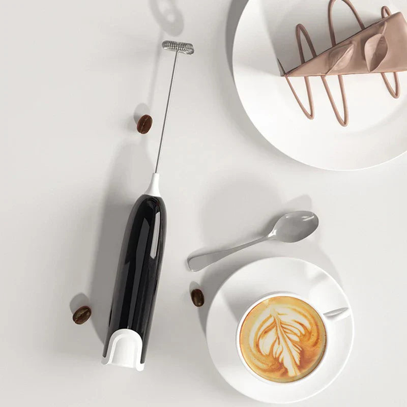 Varita espumadora - Vista superior de batidor eléctrico manual negro junto a un latte, una rebanada de pastel en un plato, una cuchara y granos de café dispersos sobre una superficie blanca