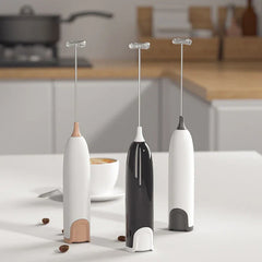 Varita espumadora - Tres batidores eléctricos manuales en blanco con rosa dorado, negro con blanco y blanco con detalles grises sobre una encimera de cocina con una taza de café al fondo