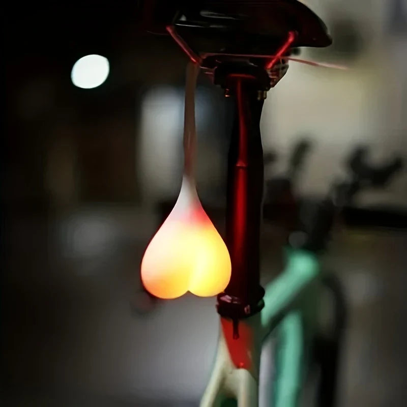 luz trasera de bicicleta con led a pilas - luz trasera de bicicleta con forma de corazón encendida colgando del sillín de una bicicleta en un entorno oscuro y desenfocado