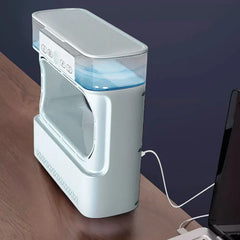 Side view of a white air cooler humidifier on a wood desk, connected to a laptop via white USB charging cable, with visible blue water in the top tank.
