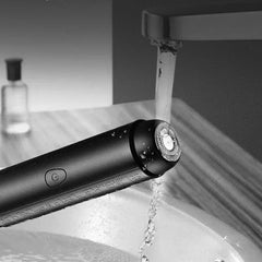 Shaving Tool - Black textured electric trimmer with circular shaving head, held under running faucet water with water droplets on its body.