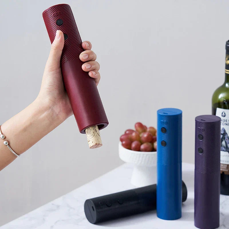 Wine Bottle Opener - A hand holding a maroon electric wine opener with a cork emerging, with three other openers and grapes in the background.