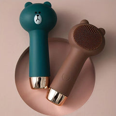Facial Cleansing Brush - Two bear-shaped silicone brushes, dark green and brown, resting on a pink circular surface
