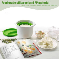 Molde para hacer queso blanco y verde sobre una mesa, junto a queso fresco, un libro de recetas, hierbas y crema de queso sazonada.