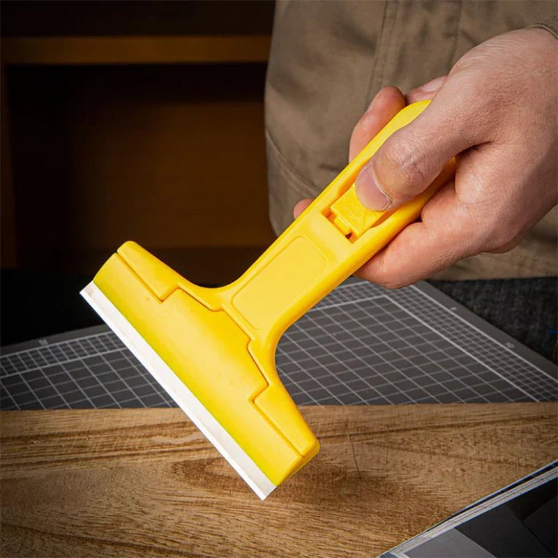 Cleaning Scraper Tool Utility Blade Remover - A hand pressing the thumb latch on a yellow scraper handle, held above a wooden surface with a cutting mat behind