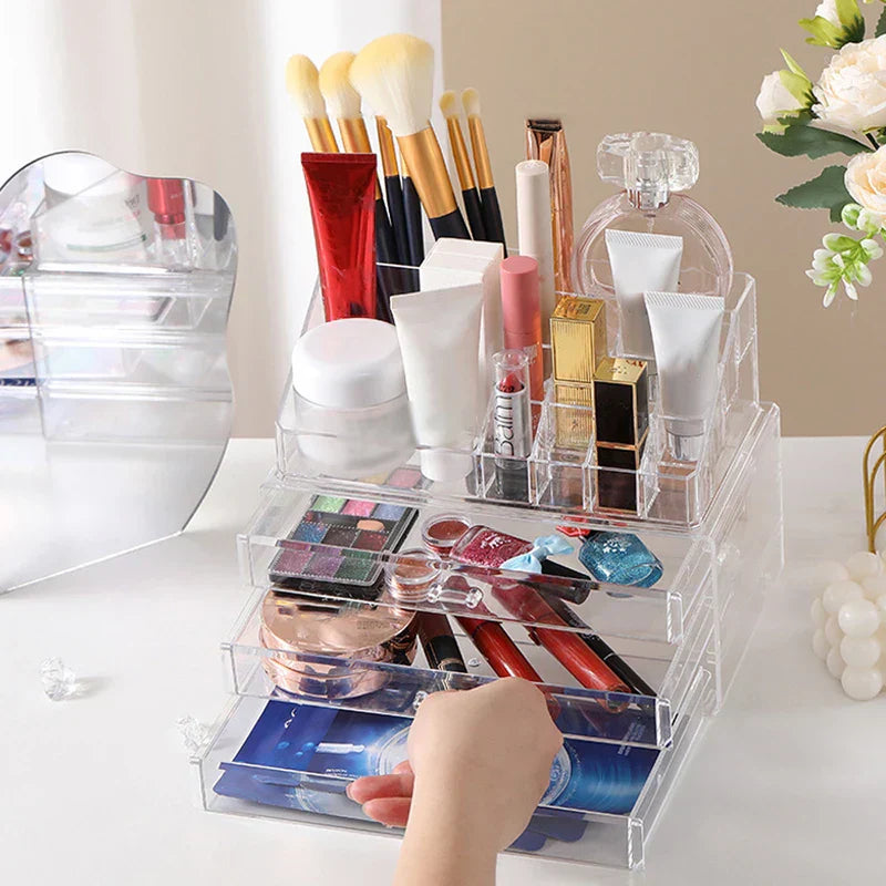 Plastic Storage Container - Clear acrylic makeup organizer with top compartments holding cosmetics, brushes, perfume, and a hand pulling out a bottom drawer with face masks.