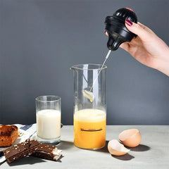 Coffee Drink Stirrer Electric Foaming Tool - Hand lifting the lid of an electric frother above a glass beaker of yellow foamy liquid, with a glass of milk, pastries, chocolate, and egg shells on the table