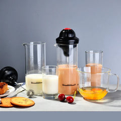 Jarras de vidrio, vasos y una taza medidora de vidrio con leche, bebida de naranja y huevos crudos, además de cerezas, galletas, un pastel y un espumador de leche sobre una superficie gris claro