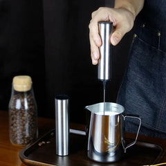 Frothing Tool - hand holding stainless steel handheld unit, frothing milk in a metal pitcher on a wooden tray with coffee beans nearby