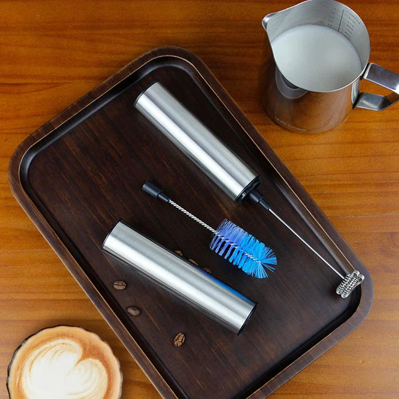 Frothing Tool - disassembled stainless steel handheld unit, cleaning brush, storage cap, and milk pitcher arranged on a wooden tray with latte art coffee