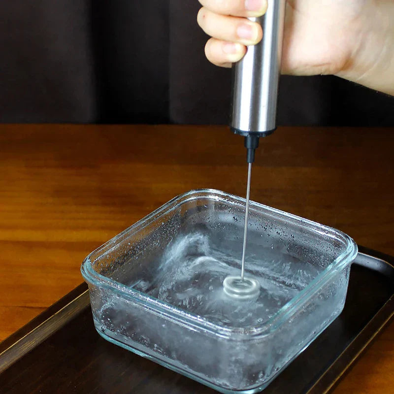 Frothing Tool - A hand holds a stainless steel handheld frother stirring clear liquid in a square glass container on a dark tray over a wooden table