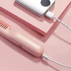 Pink heated hair brush charging via USB cable connected to a silver power bank on a pink surface