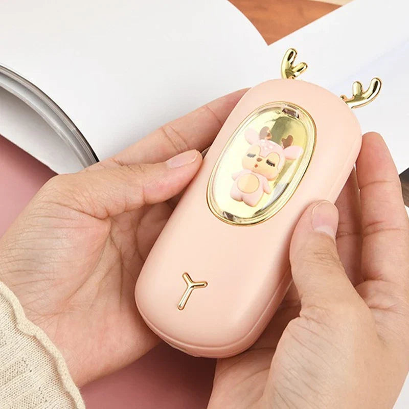 Rechargeable Hand Warmer - Two hands hold the light pink deer-designed device, with a book in the background