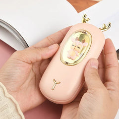 Rechargeable Hand Warmer - Two hands hold the light pink deer-designed device, with a book in the background