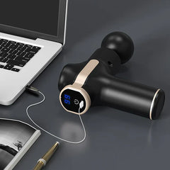 مسدس تدليك باللونين الأسود والذهبي مع شاشة رقمية مضاءة يُشحن عبر كابل USB بجانب كمبيوتر محمول ودفتر ملاحظات وقلم.