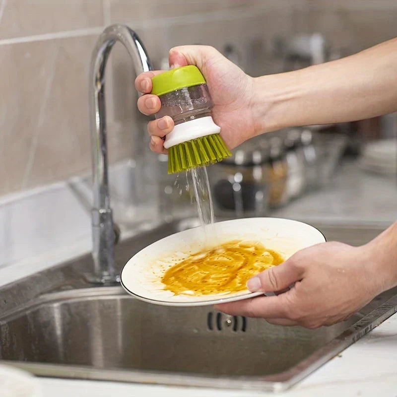 Hand pressing a green and white dish brush to release liquid while washing a sauced plate at a sink
