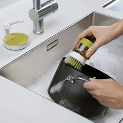 Brosse à vaisselle - Des mains utilisent la brosse avec distributeur de savon pour frotter une casserole en métal dans un évier de cuisine savonneux, la base de la brosse repose à proximité sur le comptoir