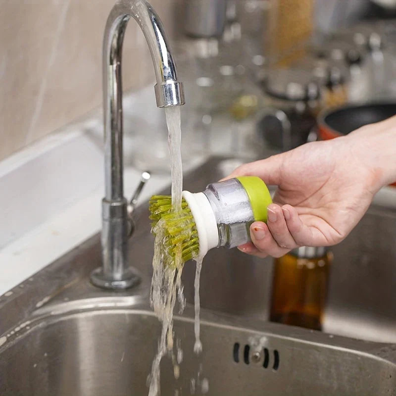 Dish Brush - A hand holds the soap dispensing brush under running faucet water to rinse it over a kitchen sink.