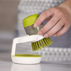 Dish Brush - A hand lifts a soap-dispensing palm brush from its matching white and green storage stand on a smooth countertop.