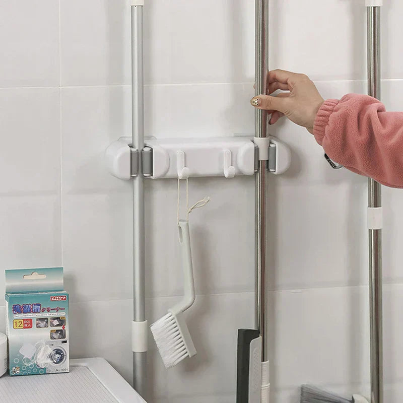 Double Mop Broom Holder - Installed white mop holder on tiled wall: two mop handles secured in slots, small cleaning brush hanging from one hook, hand touching a mop pole.