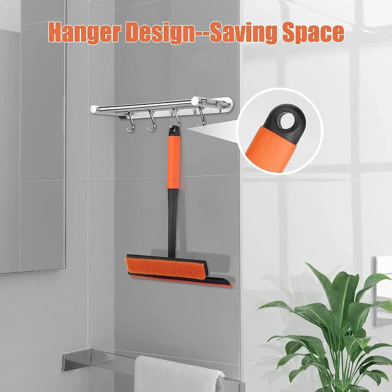 Escobilla con rasqueta: herramienta de limpieza naranja y negra colgada en un toallero cromado del baño, con un orificio para colgar en el mango, con pared de azulejos y planta de interior al fondo