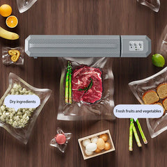 Vacuum Sealer Machine - Closed gray vacuum sealer on wood surface surrounded by vacuum-sealed dry goods, produce, meat, and loose food items