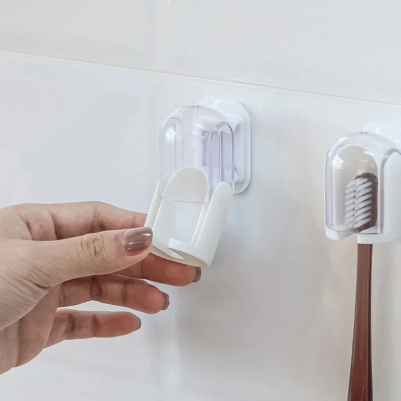 Hand pulls open the transparent cover of a wall-mounted toothbrush holder on a white tiled wall, with an identical holder holding a brown-handled toothbrush to its right.